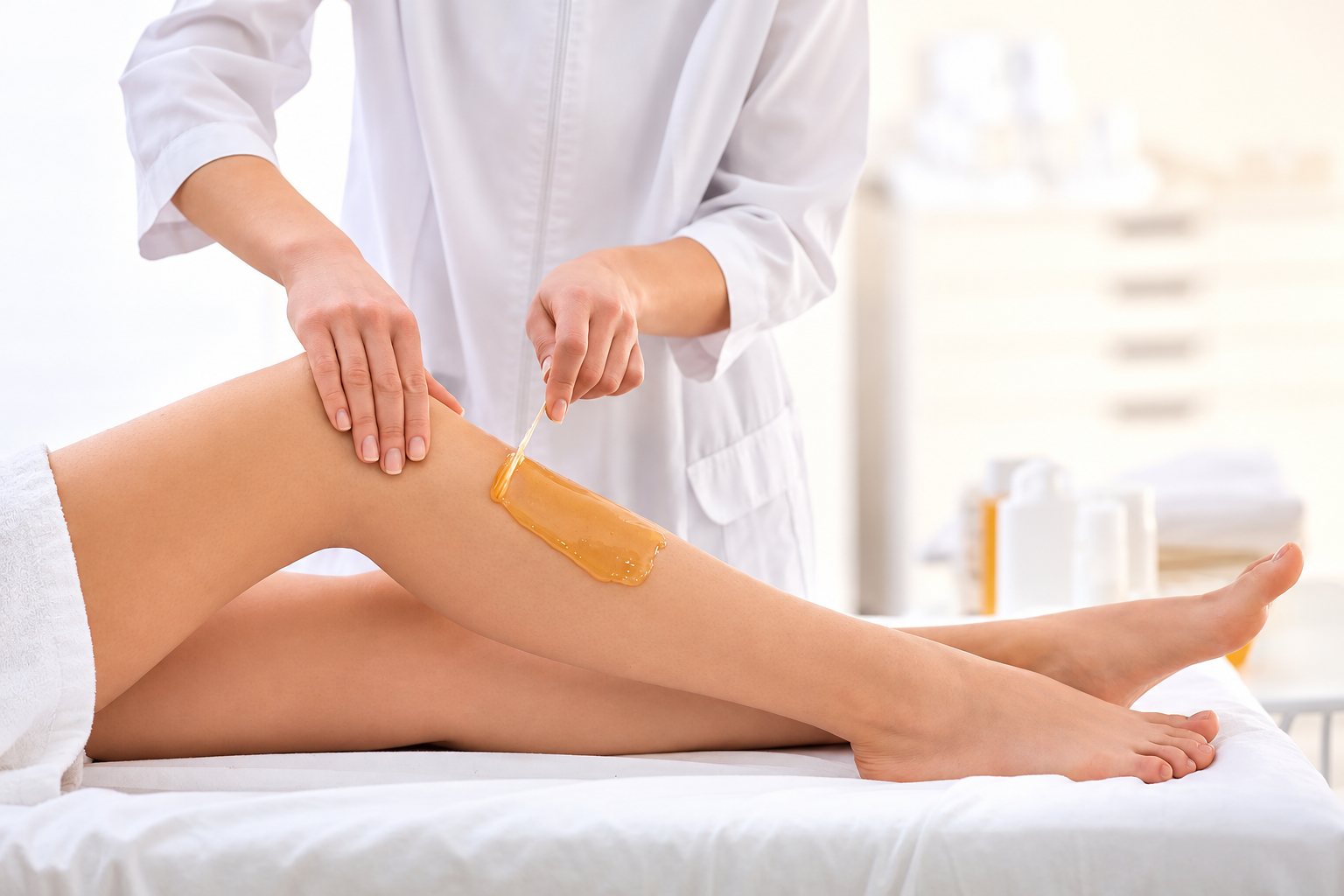 Une esthéticienne en blouse blanche étale de la cire chaude sur la jambe d'une cliente allongée sur une table de soin.