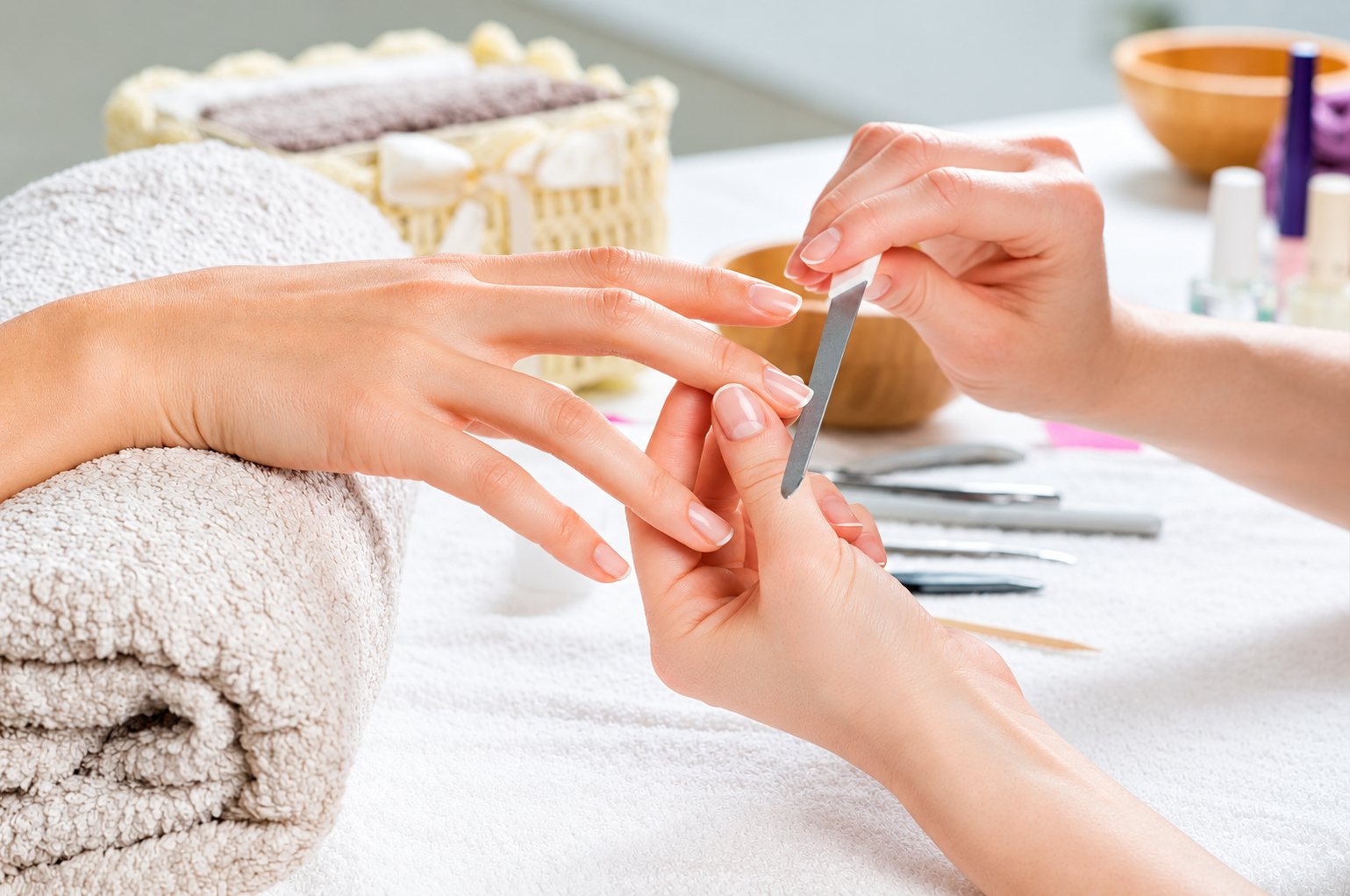 Une manucure limant les ongles d'une cliente, la main posée sur une serviette roulée, avec des outils et des bols en bois autour.