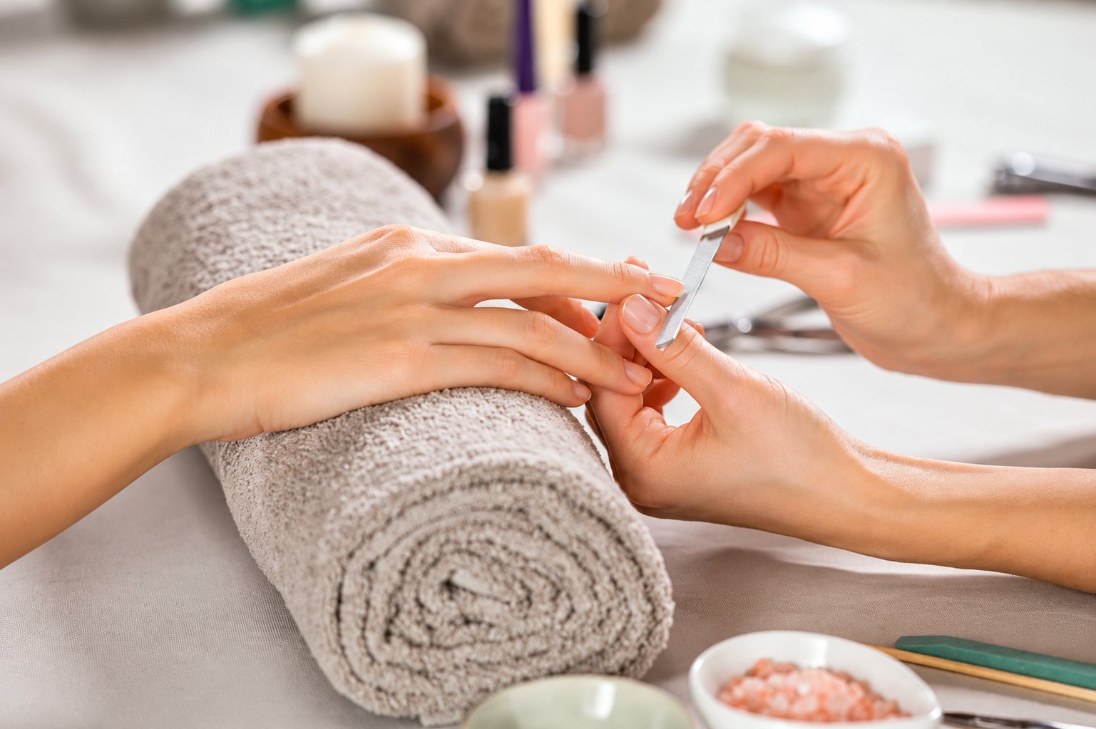 Une manucure lime les ongles d'une cliente dont la main repose sur une serviette beige roulée, avec des vernis et un bol de sel rose en arrière-plan.