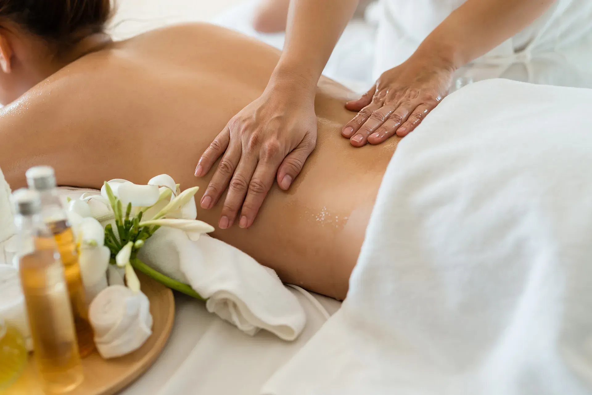 Une masseuse applique de l'huile sur le dos d'une cliente allongée, avec des flacons d'huile, des serviettes blanches et des fleurs sur un plateau en bois au premier plan.
