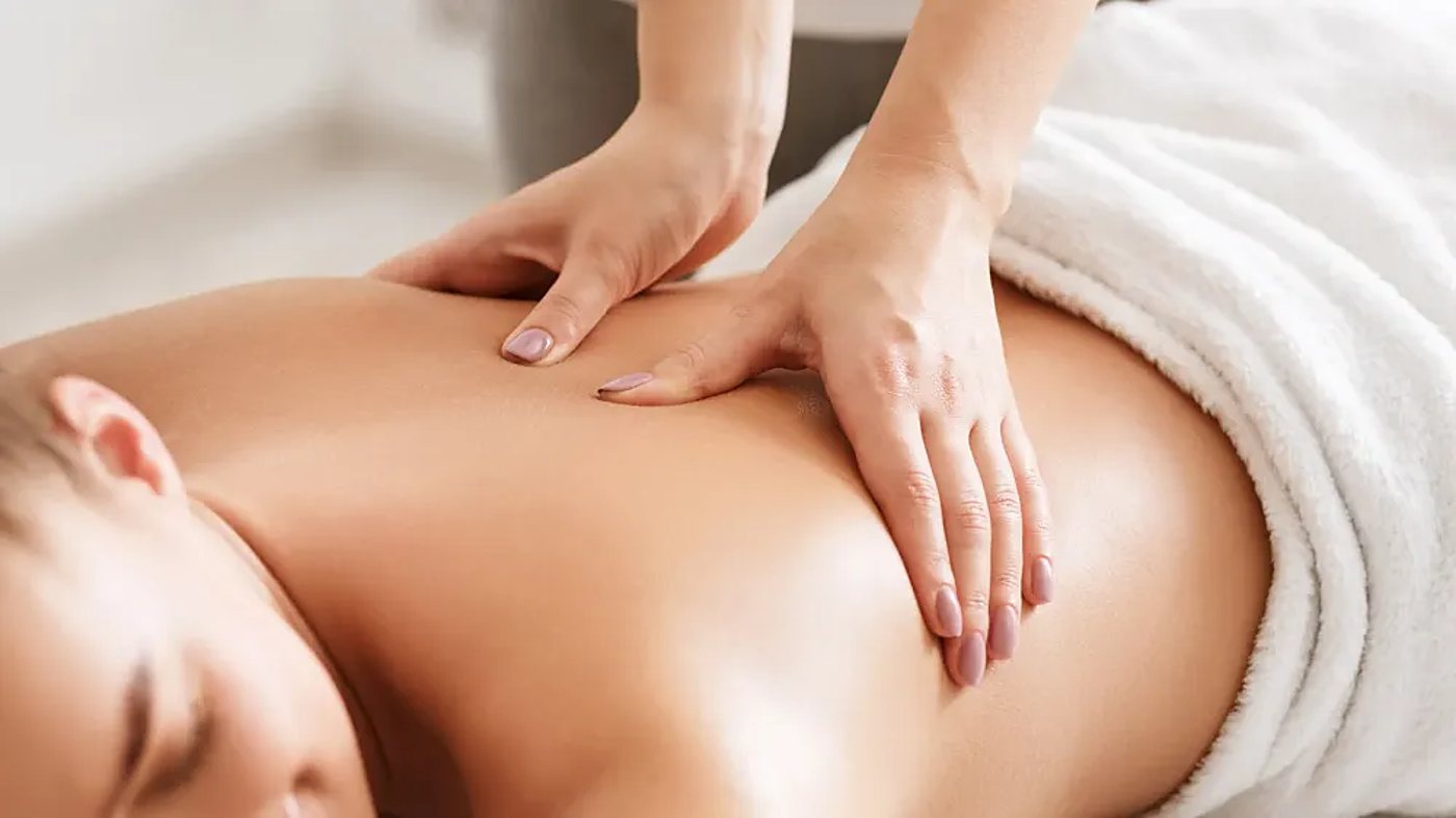 Une praticienne aux ongles vernis masse le dos d'une femme allongée, une serviette blanche posée sur elle.