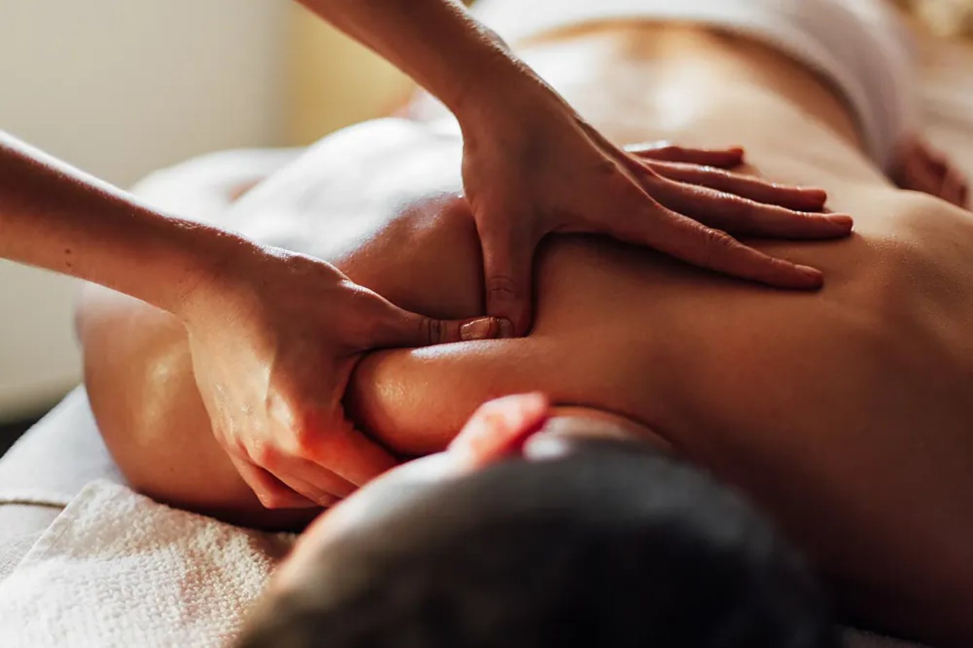 Les mains d'une praticienne massent l'épaule et le dos d'une personne allongée sur une table de massage recouverte d'une serviette blanche.