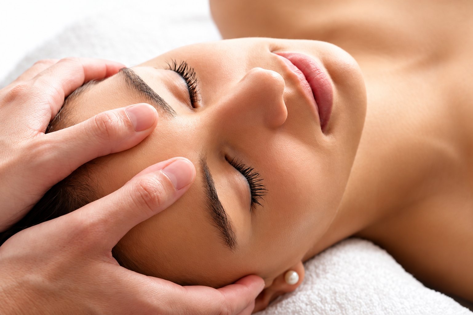 Une femme allongée sur une serviette blanche, les yeux fermés, reçoit un massage du visage et du front par deux mains.