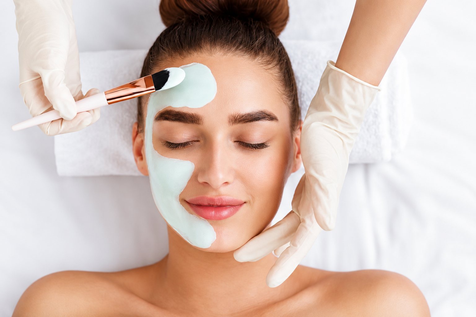Une jeune femme allongée, les yeux fermés, reçoit l'application d'un masque vert sur le visage au pinceau par une esthéticienne gantée.